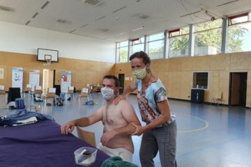 Eine Frau mit Mund- und Nasenschutz behandelt die Schulter eines Seminarteilnehmers